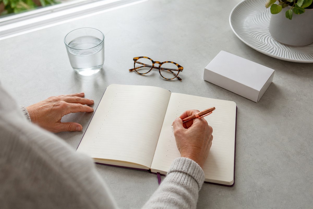 Handen schrijven vragen in een notitieboek naast een bril en een glas water